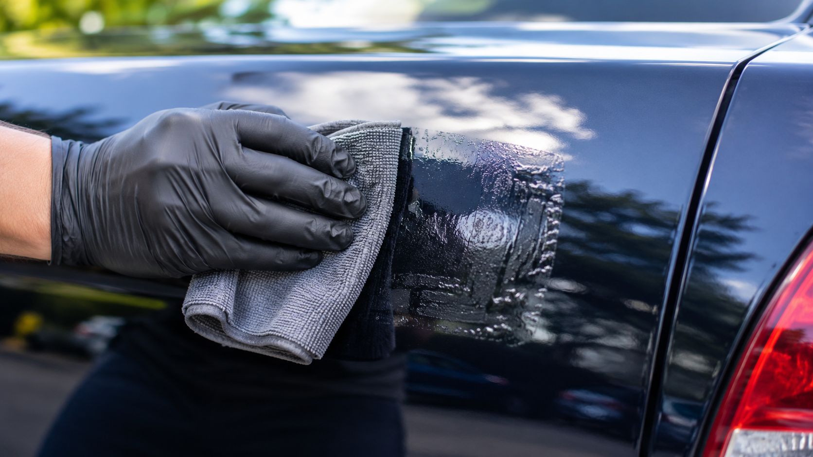 A person wearing a black glove uses a gray microfiber cloth to remove a decal from a car.